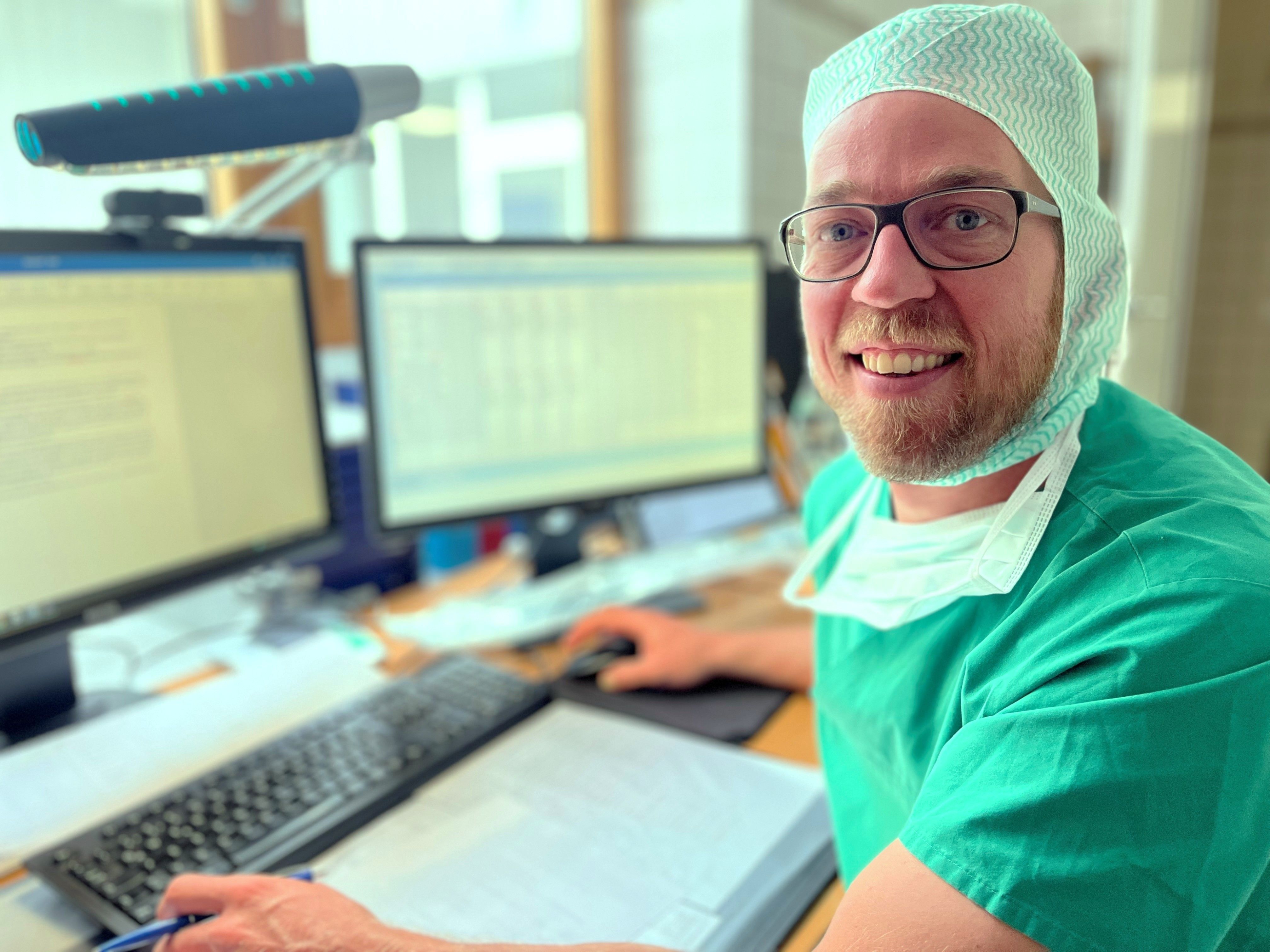 Patrick Dorsten sitzt in OP-Kleidung am PC und lächelt in die Kamera. 