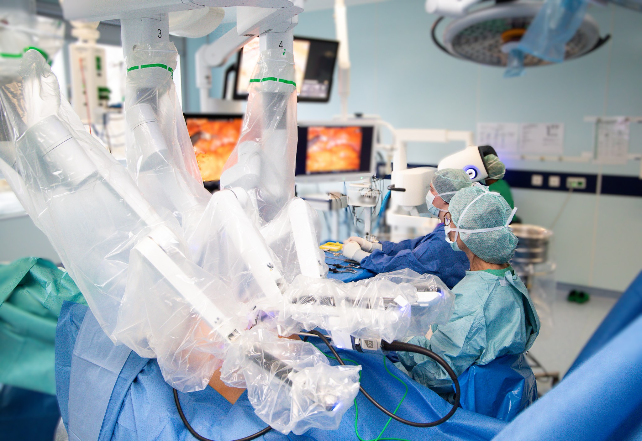 Blick in den OP, Roboterarme arbeiten im Patienten, Operateurin mit Assistentin am Tisch, in Hintergrund Operateurin an der Konsole. 