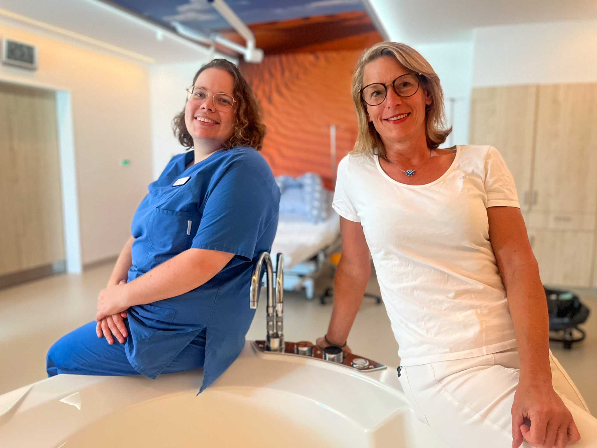 Wiebke Zimmermann und Frauke Sonntag sitzen auf einem Geburtsbett im Kreißsaal und lächeln in die Kamera. 