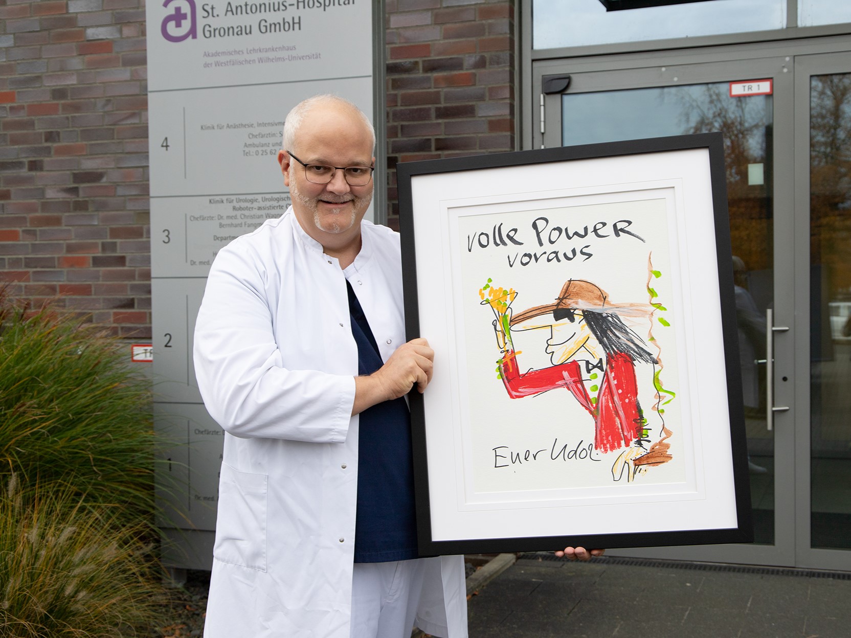 Chefarzt Dr. Christian Wagner steht mit dem Gemälde von Udo Lindenberg im Arm vor dem Eingang des Krankenhauses und lächelt in die Kamera.  