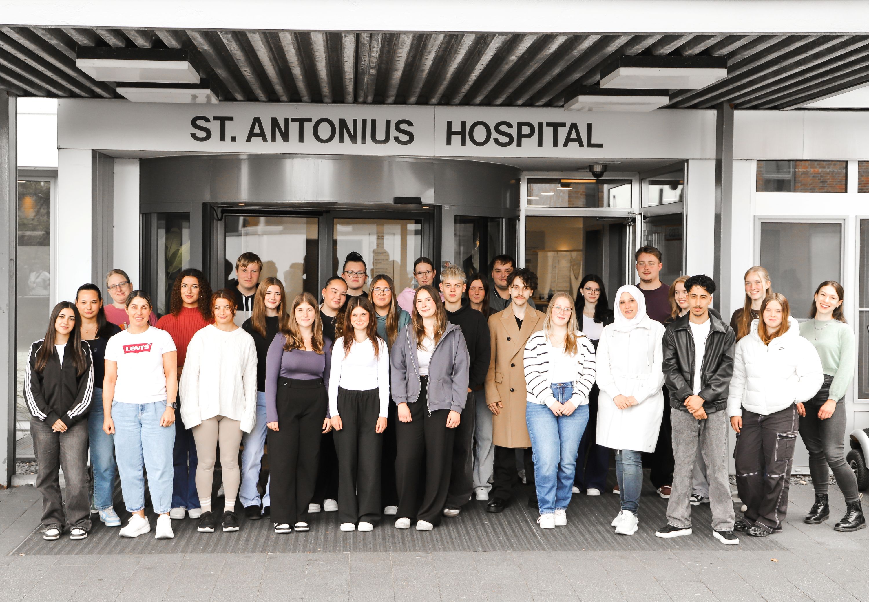 Alle Auszubildenden haben sich vor den Eingang des St. Antonius-Hospitals Gronau postiert und schauen in die Kamera. 
