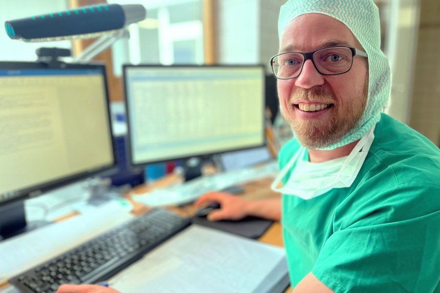 Patrick Dorsten sitzt in OP-Kleidung am PC und lächelt in die Kamera. 