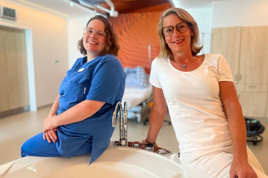 Wiebke Zimmermann und Frauke Sonntag sitzen auf einem Geburtsbett im Kreißsaal und lächeln in die Kamera. 