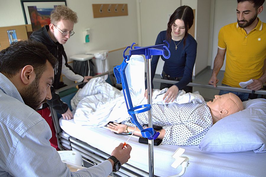 Pflegeauszubildende stehen um ein Pflegebett mit einer Patientenpuppe herum und suchen nach Fehlern, die gemacht wurden. 