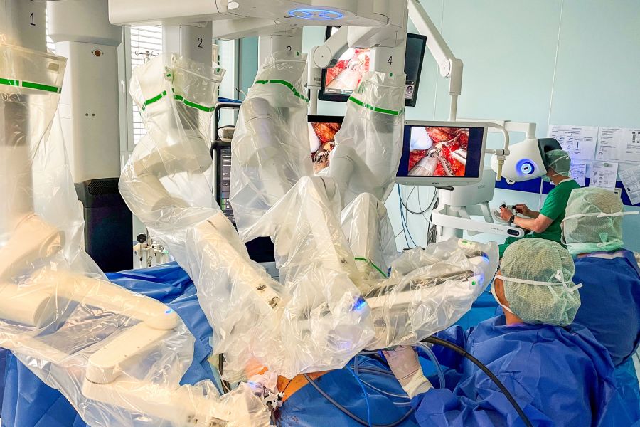 Ärzte im OP stehen am Tisch mit den Armen des neuen da Vinci 5 Roboter-Systems. Im Hintergrund der operierende Arzt an der Konsole. 