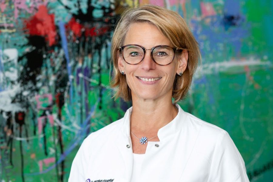 Chefärztin Frauke Sonntag im weißen Kittel vor buntem Gemälde.