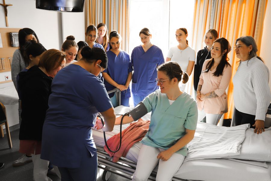 Eine Schülerin misst bei einer anderen den Blutdruck, während die Gruppe von Schülerinnen und Lehrerinnen der Pflegeschule um das Krankenbett herumstehen. 
