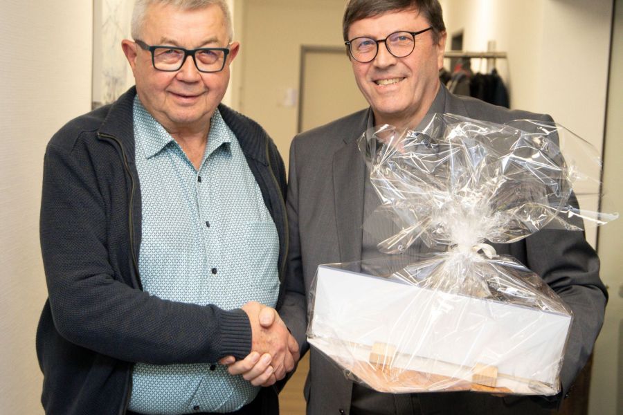 Bernward Wigger (rechts) überreicht Ludger Berges ein Geschenk, beide geben sich die Hand und lächeln in die Kamera. 