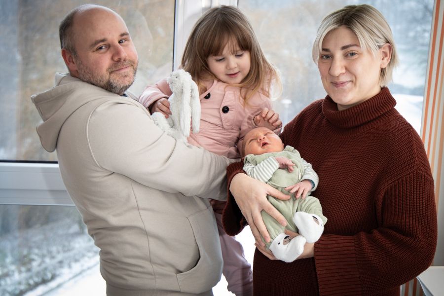 Vater mit älterer Tochter im Arm und Mutter mit dem neugeborenen Baby im Arm stehen vor dem Fenster des Krankenhauses und lächeln in die Kamera. 