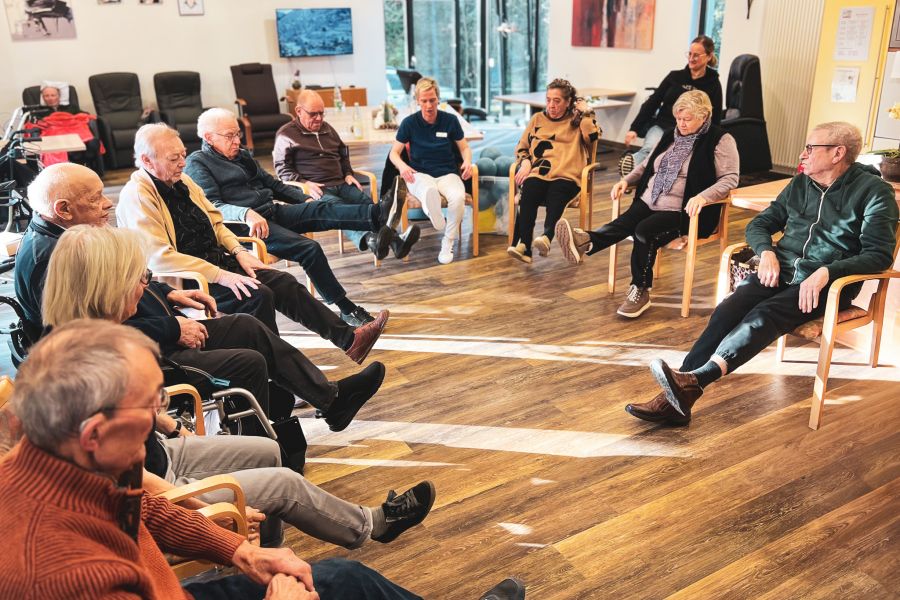 Senioren sitzen im Stuhlreis und halten ein Bein gestreckt für eine gemeinsame Gymnastikübung. 