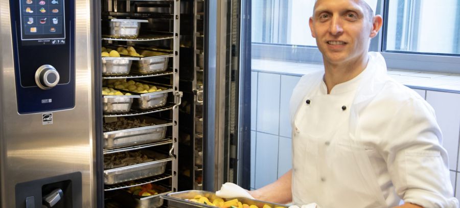 Koch steht mit einem Blech frisch zubereitetem Essen vor dem Garofen. 