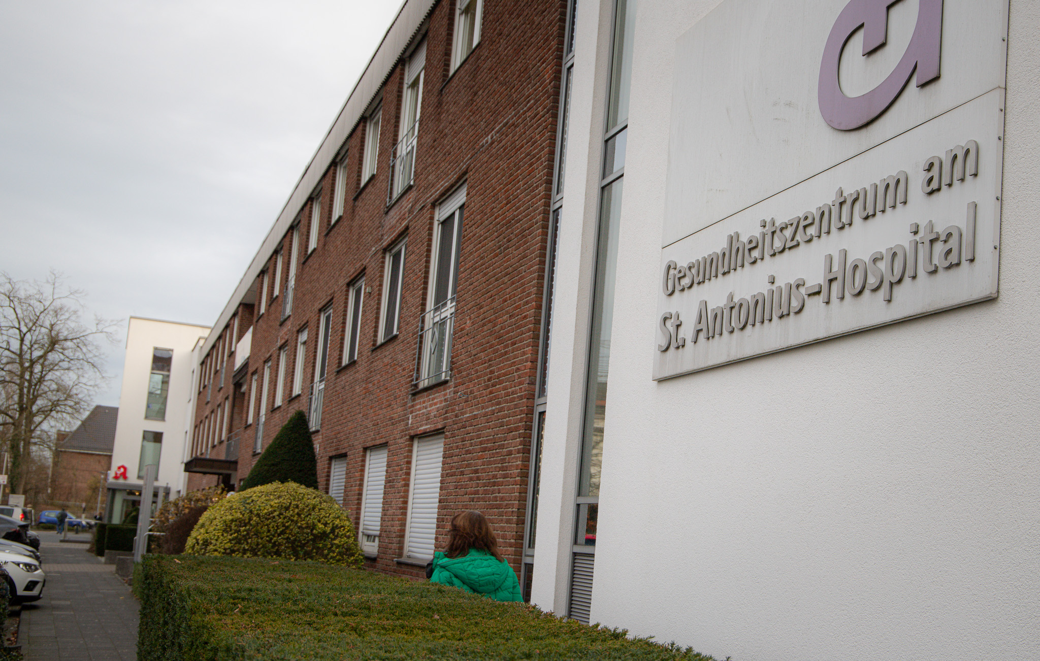 Die kardiologische Praxis im Gesundheitszentrum des St. Antonius-Hospitals Gronau zieht innerhalb des Gebäudes am Möllenweg 22 um. Blick auf das Gebäude des MVZ mit Schild Gesundheitszentrum im Vordergrund und einer Frau, die den Weg entlang läuft (von hinten). .